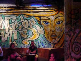 A vibrant graffiti mural covers a wall, featuring a large, stylized face with striking blue eyes and intricate patterns. The background includes abstract shapes and graffiti writing. Several people are seated at tables in front, creating a casual and social atmosphere.
