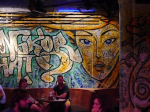 A vibrant graffiti mural covers a wall, featuring a large, stylized face with striking blue eyes and intricate patterns. The background includes abstract shapes and graffiti writing. Several people are seated at tables in front, creating a casual and social atmosphere.
