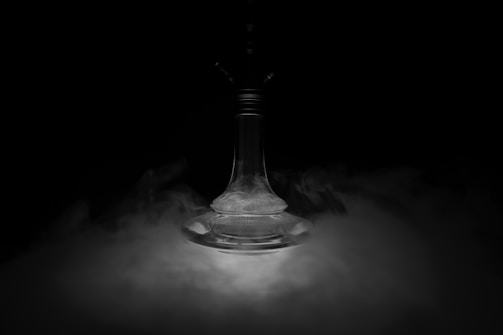 A glass hookah base is surrounded by swirling smoke against a dark background. The lighting creates an ethereal atmosphere, highlighting the curves and texture of the glass.