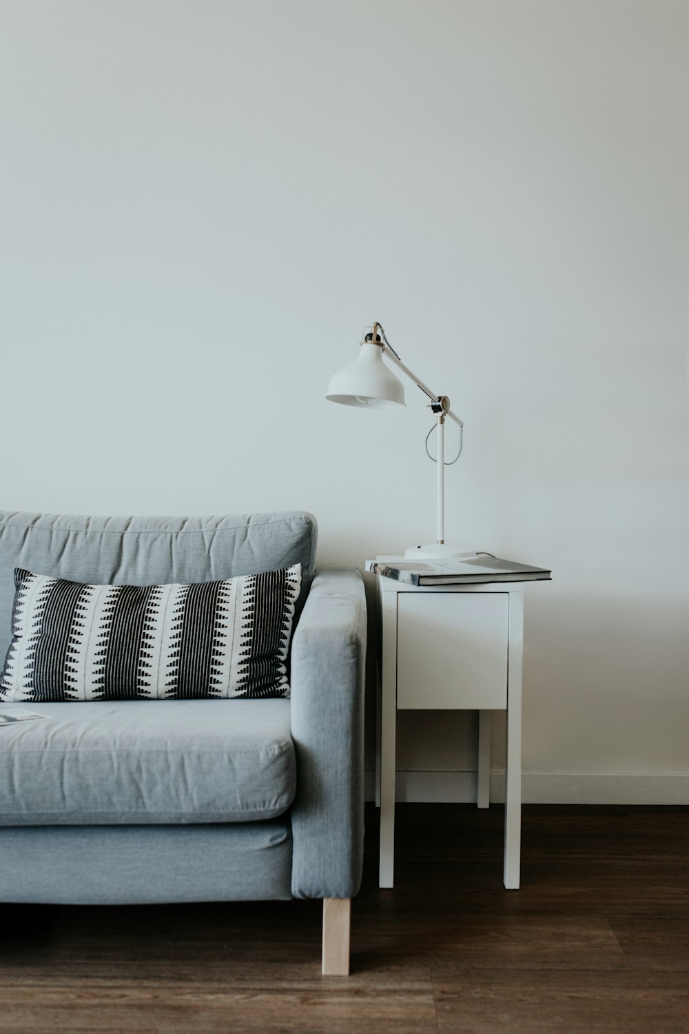 Lámpara de estudio blanca en la parte superior de la mesa auxiliar de madera blanca junto al sofá de tela gris