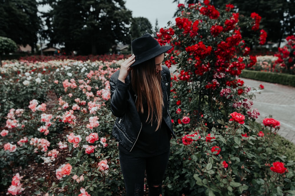 mulher na jaqueta preta do terno e chapéu perto do campo de rosas rosa e vermelhas