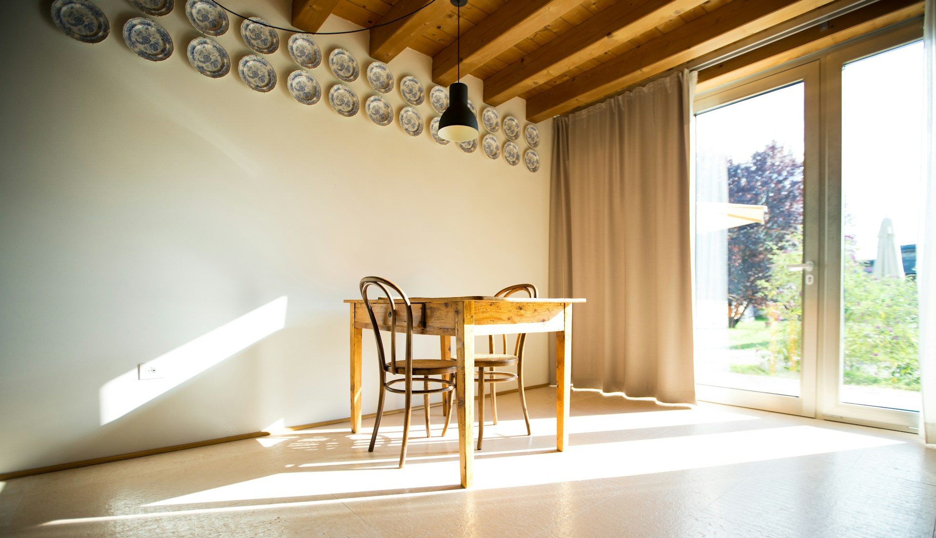 A minimalist dining area bathed in natural light, showcasing a sleek white table set with neutral-toned tableware and surrounded by light wood chairs with clean lines.