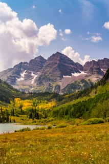 A breathtaking view of rugged mountains with distinct peaks, partially covered in snow. In the foreground, a vibrant valley filled with lush greenery and patches of yellow foliage frames a serene lake. The sky is bright with scattered fluffy clouds, enhancing the natural beauty of the landscape.