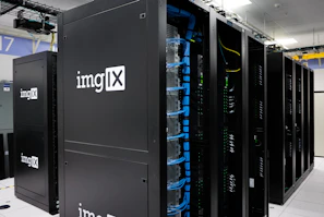 Several server racks with the logo 'imgIX' are neatly arranged in a data center. The racks are equipped with blue and yellow cable management, ensuring proper organization of the cables inside. The room is well-lit with overhead lighting and has a modern, clean appearance.