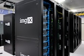 Several server racks with the logo 'imgIX' are neatly arranged in a data center. The racks are equipped with blue and yellow cable management, ensuring proper organization of the cables inside. The room is well-lit with overhead lighting and has a modern, clean appearance.