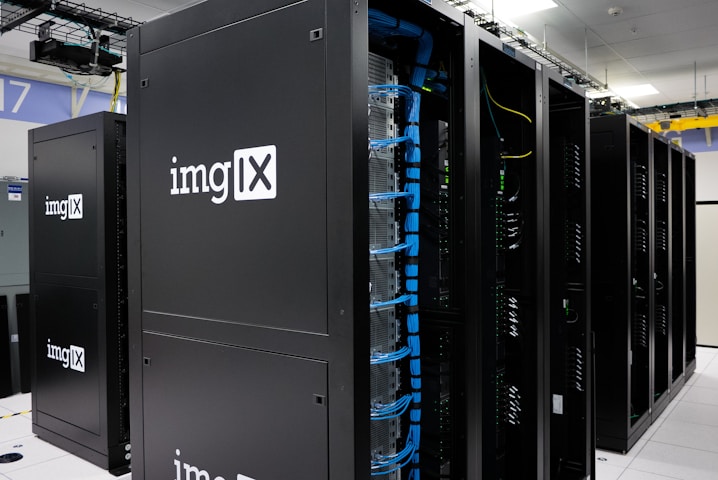 Several server racks with the logo 'imgIX' are neatly arranged in a data center. The racks are equipped with blue and yellow cable management, ensuring proper organization of the cables inside. The room is well-lit with overhead lighting and has a modern, clean appearance.
