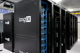Several server racks with the logo 'imgIX' are neatly arranged in a data center. The racks are equipped with blue and yellow cable management, ensuring proper organization of the cables inside. The room is well-lit with overhead lighting and has a modern, clean appearance.
