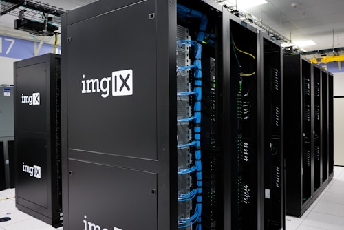 Several server racks with the logo 'imgIX' are neatly arranged in a data center. The racks are equipped with blue and yellow cable management, ensuring proper organization of the cables inside. The room is well-lit with overhead lighting and has a modern, clean appearance.