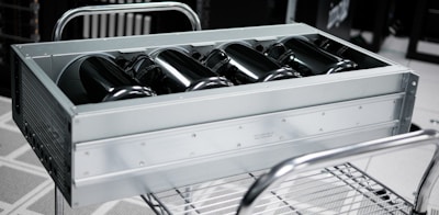 A metallic server rack is positioned on a wheeled cart. The server chassis is open, revealing black cylindrical components arranged in a row inside. The background is blurred, indicating a data center environment with additional server racks.