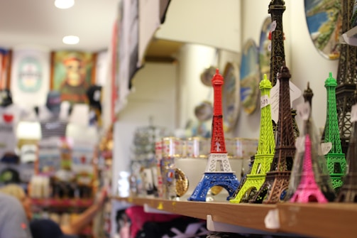 Souvenir shop display with miniature Eiffel Tower replicas in various colors, including red, blue, green, and pink. The background shows shelves filled with other merchandise, blurred to emphasize the foreground objects.