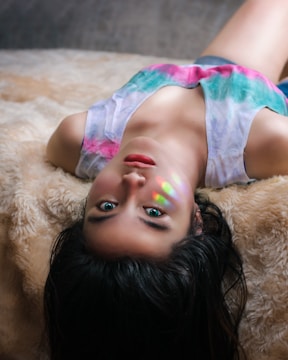 A person lying on a soft, textured surface, with long dark hair spread out. The face is illuminated by a small spectrum of rainbow colors across the cheek. The individual's eyes are open and looking towards the camera, wearing a sleeveless top with a blend of pink and green colors.