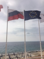 Three flags are seen waving in the breeze on tall poles. Behind them, the sea stretches out under a cloudy sky, bordered by a rocky shore and beach umbrellas lined up along the coast.