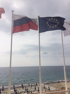 Three flags are seen waving in the breeze on tall poles. Behind them, the sea stretches out under a cloudy sky, bordered by a rocky shore and beach umbrellas lined up along the coast.