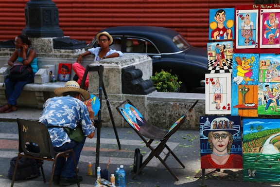 A street artist wearing a straw hat and a blue floral shirt paints at an outdoor setup, with an array of vibrant, colorful paintings displayed on wooden frames nearby. The scene is set against a background that includes a vintage black car and a red wall. A man in a hat and sunglasses relaxes against a stone barrier nearby, while a woman sits on a bench using her phone.