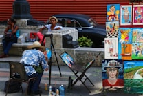 A street artist wearing a straw hat and a blue floral shirt paints at an outdoor setup, with an array of vibrant, colorful paintings displayed on wooden frames nearby. The scene is set against a background that includes a vintage black car and a red wall. A man in a hat and sunglasses relaxes against a stone barrier nearby, while a woman sits on a bench using her phone.