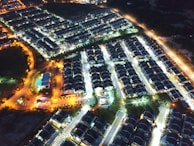 Aerial view of a residential neighborhood at night, with streets illuminated by streetlights. Numerous houses are neatly aligned in rows, creating a grid-like pattern. Some areas are brightly lit with white lights while others have a warm yellow glow. A few larger buildings and open spaces are visible, and the surrounding area appears darker with less lighting.