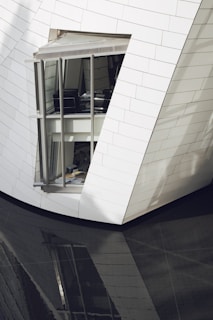 A modern architectural structure with sleek, angled walls clad in white tiles. A large window reveals an interior space with office furniture. The building's reflection is visible on a glossy, dark surface below.