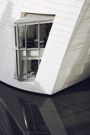 A modern architectural structure with sleek, angled walls clad in white tiles. A large window reveals an interior space with office furniture. The building's reflection is visible on a glossy, dark surface below.