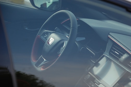 A view of a car's interior through the window, showcasing a steering wheel with the Honda logo prominently displayed. The dashboard includes a digital display and various controls.