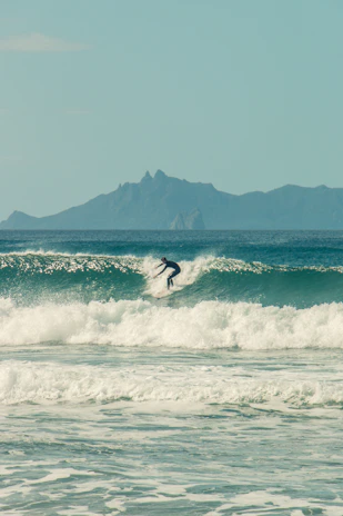 A smiling guest catching a wave on a surfboard with volcanic mountains in the background under a clear blue sky.