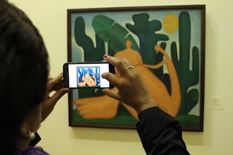 A person is taking a photo with a smartphone of a colorful painting featuring abstract shapes and a reclining figure. The art piece has a mixture of organic shapes that resemble cacti and large leaves, with a sun in the background.