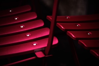 A close-up view of red, slatted metal chairs with rounded edges, featuring visible rivets. The lighting creates shadows and highlights that emphasize the texture and color of the chairs, set against a dark background.