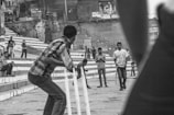 A group of people are engaged in a street cricket game. The scene captures a batsman in the foreground preparing for a delivery, while other individuals observe and participate in the game. The backdrop features a series of steps and buildings with graffiti, suggesting an urban environment.