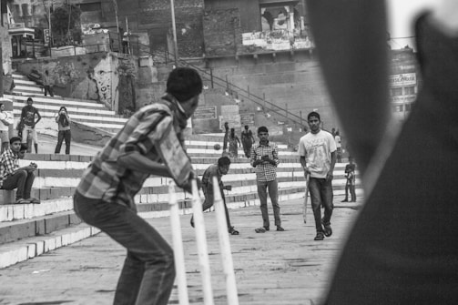 A group of people are engaged in a street cricket game. The scene captures a batsman in the foreground preparing for a delivery, while other individuals observe and participate in the game. The backdrop features a series of steps and buildings with graffiti, suggesting an urban environment.