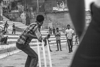 A group of people are engaged in a street cricket game. The scene captures a batsman in the foreground preparing for a delivery, while other individuals observe and participate in the game. The backdrop features a series of steps and buildings with graffiti, suggesting an urban environment.