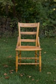 A wooden chair with intricate carvings sits on lush green grass, surrounded by a backdrop of dense foliage. The chair's design is traditional, showcasing vertical slats on the backrest and a sturdy build.