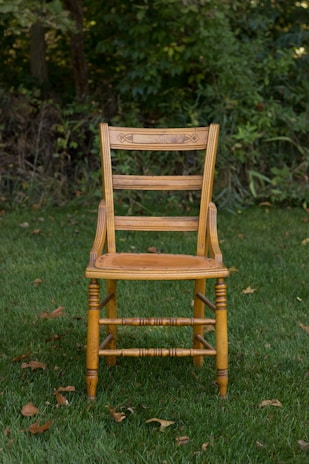 A wooden chair with intricate carvings sits on lush green grass, surrounded by a backdrop of dense foliage. The chair's design is traditional, showcasing vertical slats on the backrest and a sturdy build.