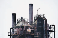 Industrial structures featuring large, cylindrical towers and complex piping systems. The surfaces appear aged and rusted, with graffiti visible on one of the towers. Metal railings and ladders are part of the construction.