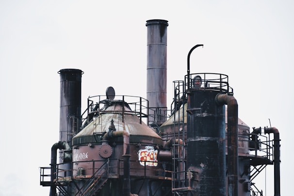Industrial structures featuring large, cylindrical towers and complex piping systems. The surfaces appear aged and rusted, with graffiti visible on one of the towers. Metal railings and ladders are part of the construction.