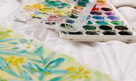 A palette of watercolors with various shades is resting on a white surface next to a floral watercolor painting featuring green leaves and yellow flowers.