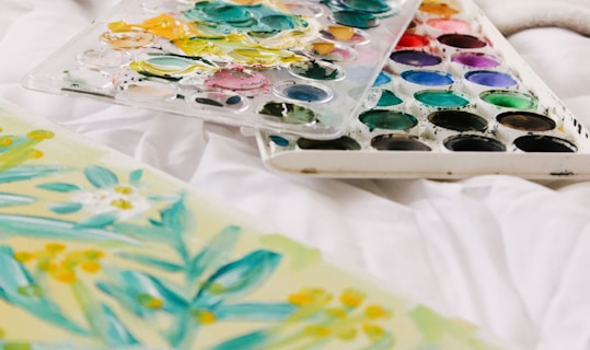 A palette of watercolors with various shades is resting on a white surface next to a floral watercolor painting featuring green leaves and yellow flowers.