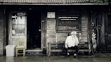 A rustic wooden storefront with a weathered appearance features signs indicating a shoe repair shop and a cultural center. An older man with white hair sits thoughtfully on a wooden bench outside, creating a scene that evokes a sense of nostalgia. The entrance displays a wooden chair and a makeshift trash bin, surrounded by ivy creeping up the wall.
