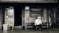 A rustic wooden storefront with a weathered appearance features signs indicating a shoe repair shop and a cultural center. An older man with white hair sits thoughtfully on a wooden bench outside, creating a scene that evokes a sense of nostalgia. The entrance displays a wooden chair and a makeshift trash bin, surrounded by ivy creeping up the wall.