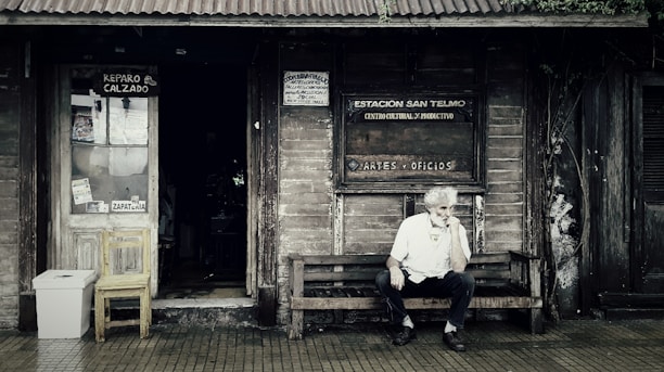 A rustic wooden storefront with a weathered appearance features signs indicating a shoe repair shop and a cultural center. An older man with white hair sits thoughtfully on a wooden bench outside, creating a scene that evokes a sense of nostalgia. The entrance displays a wooden chair and a makeshift trash bin, surrounded by ivy creeping up the wall.