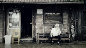 A rustic wooden storefront with a weathered appearance features signs indicating a shoe repair shop and a cultural center. An older man with white hair sits thoughtfully on a wooden bench outside, creating a scene that evokes a sense of nostalgia. The entrance displays a wooden chair and a makeshift trash bin, surrounded by ivy creeping up the wall.