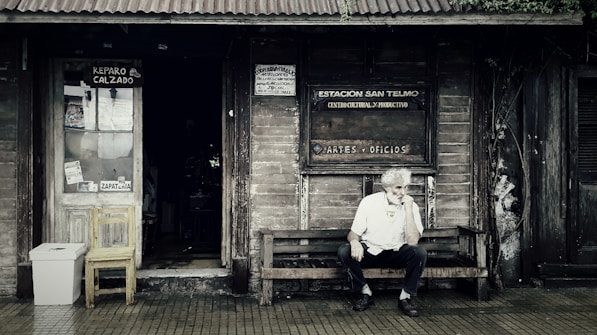 A rustic wooden storefront with a weathered appearance features signs indicating a shoe repair shop and a cultural center. An older man with white hair sits thoughtfully on a wooden bench outside, creating a scene that evokes a sense of nostalgia. The entrance displays a wooden chair and a makeshift trash bin, surrounded by ivy creeping up the wall.