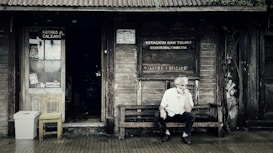 A rustic wooden storefront with a weathered appearance features signs indicating a shoe repair shop and a cultural center. An older man with white hair sits thoughtfully on a wooden bench outside, creating a scene that evokes a sense of nostalgia. The entrance displays a wooden chair and a makeshift trash bin, surrounded by ivy creeping up the wall.