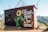 A small brown utility building decorated with a colorful mural of a sunflower and green leaves. The words 'Always Room to Grow' are painted in large white script. Electrical wires and equipment are visible, and the building is surrounded by a natural landscape with hills and greenery.