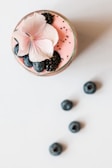 A pink smoothie in a glass is topped with blackberries, blueberries, chia seeds, and a light pink flower. Several blueberries are scattered on the white surface next to the glass.