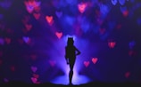 A silhouette of a person with cat ears stands against a dark purple and blue background with bokeh heart shapes in red and pink. The person is positioned in the center with a confident stance.