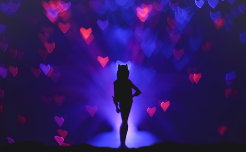 A silhouette of a person with cat ears stands against a dark purple and blue background with bokeh heart shapes in red and pink. The person is positioned in the center with a confident stance.