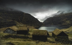 A dramatic landscape featuring rustic wooden houses with grass roofs nestled in a verdant valley. Surrounding the homes are steep, rugged hillsides under a thick layer of dark, ominous clouds. A calm body of water is visible in the mid-ground, bordered by the majestic, mist-covered mountains.