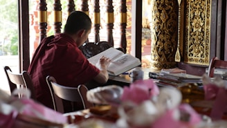 A person dressed in a maroon robe is sitting at a wooden table, focused on reading a large open book. The setting appears to be a study area or library, with stacks of books and a cup nearby. Intricately carved golden wooden decorations and a series of wooden pillars are visible in the background, adding to a traditional and serene atmosphere.