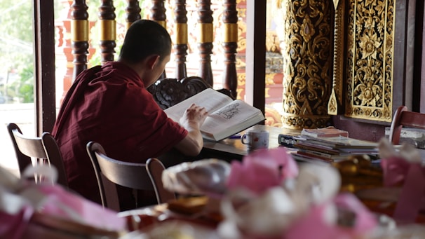 A person dressed in a maroon robe is sitting at a wooden table, focused on reading a large open book. The setting appears to be a study area or library, with stacks of books and a cup nearby. Intricately carved golden wooden decorations and a series of wooden pillars are visible in the background, adding to a traditional and serene atmosphere.
