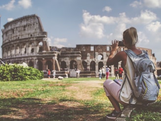 A person wearing a backpack is sitting on the grass, taking a photo of the Colosseum in Rome. The ancient amphitheater is partially visible, with tourists walking around. The scene is bright with a partly cloudy sky.