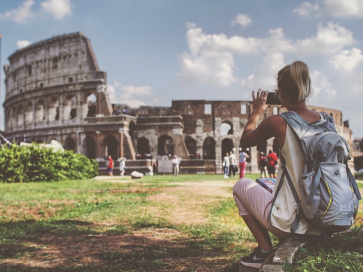 A person wearing a backpack is sitting on the grass, taking a photo of the Colosseum in Rome. The ancient amphitheater is partially visible, with tourists walking around. The scene is bright with a partly cloudy sky.